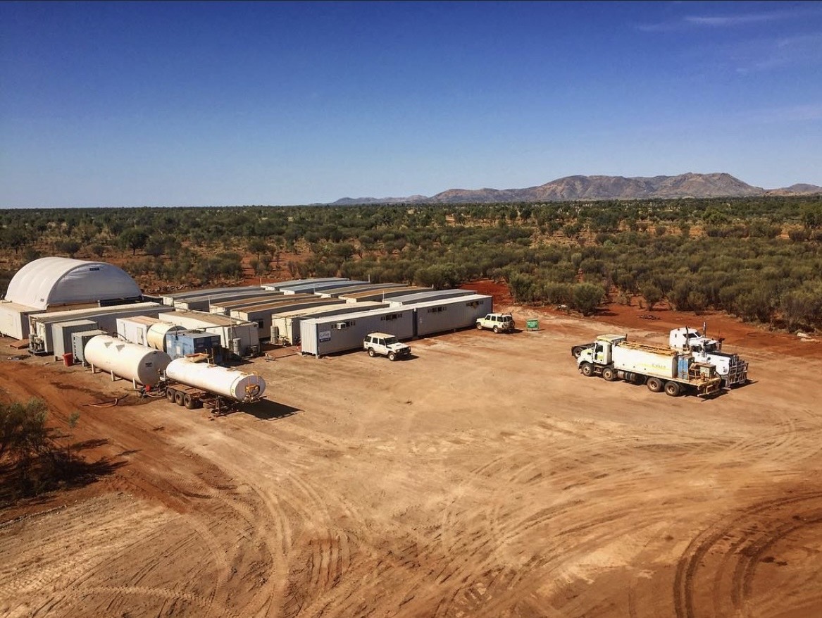 Aerial view of a remote construction camp with modular buildings, fuel tanks, and service trucks on red earth